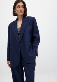 Blazer ajusté bleu marine avec un bouton unique, des revers nets et une poche poitrine unique, associé à un pantalon ample assorti. Texture lisse.