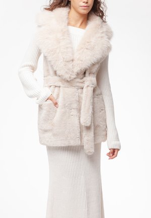 Donna che indossa un vestito bianco in maglia a maniche lunghe sotto un gilet beige in ecopelliccia con cintura e ampio colletto, con la mano in tasca.