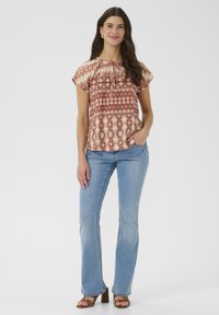 Bedruckte Bluse in Rot und Beige mit Rundhalsausschnitt und kurzen, schulterfreien Ärmeln. Kombiniert mit hellblauen Schlagjeans und braunen Absatzsandalen.