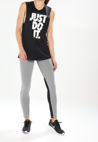 Sort ærmeløs atletisk tanktop med hvid "JUST DO IT." tekst, parret med grå leggings med sorte sidepaneler og sorte atletiske sko.
