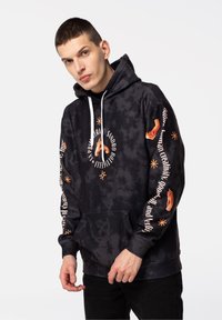 Crna dukserica s tie-dye uzorkom, koja ima prednji džep, bijele uzice i narančaste grafičke otiske, uključujući ruke i tekst duž rukava.