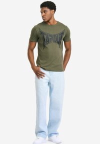 Olivgrünes Baumwoll-T-Shirt mit einem großen schwarzen "TAPOUT"-Logo, kombiniert mit hellblauen Denim-Jeans und weißen Turnschuhen.