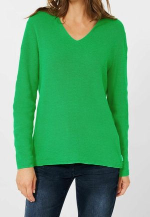 Femme portant un pull en tricot vert vif à col en V avec des manches longues et un jean bleu foncé, montrée des épaules aux hanches.