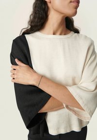 Blouse color block noir et crème avec des manches larges, présentant un tissu texturé et un ourlet arrondi. Détail d'une main reposant sur le bras.