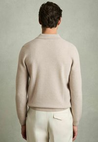 Pull en tricot beige clair, avec un col, silhouette ajustée et manches longues, porté avec un pantalon de couleur claire. Texture douce, détails minimalistes.