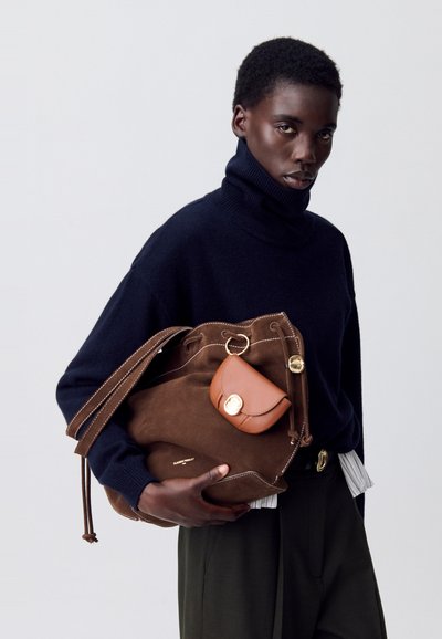 Borsa a secchiello in suede marrone con una pochette più piccola color cammello attaccata, dotata di dettagli in oro. Modella con dolcevita blu navy, pantaloni verdi e dettagli a righe bianche.