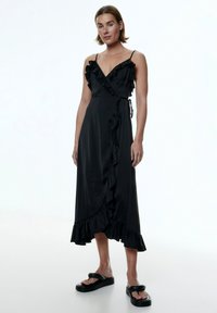 Schwarzes Satin-Wickelkleid mit Rüschensaum, verstellbaren Trägern und einem ausgestellten Saum. Kombiniert mit schwarzen Plattform-Sandalen mit minimalistischem Design.