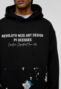 Zwarte hoodie met witte tekst: "REVOLUTIONIZE ART DESIGN PROCESSES" en spetters van blauwe en grijze verf aan de onderkant. Met trekkoordkap.