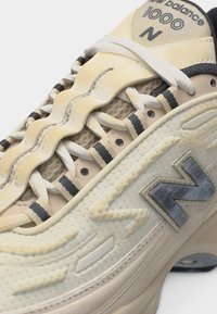 Gros plan sur une basket New Balance 1000 N beige montrant la tige texturée, le protège-lacets ondulé, les lacets blancs et le logo "N" cousu sur le côté.