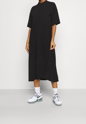 Kvinde iført en sort midi-kjole med knapper og korte ærmer samt hvide Nike-sneakers med hvide sokker, stående på en grå gulv.