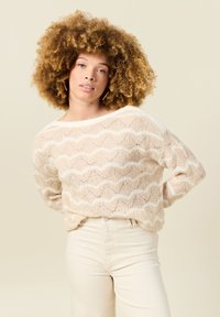 Gebreid beige sweater met witte golfpatroon, ronde halslijn en lange mouwen. Gecombineerd met lichtgekleurde, slim-fit broek.