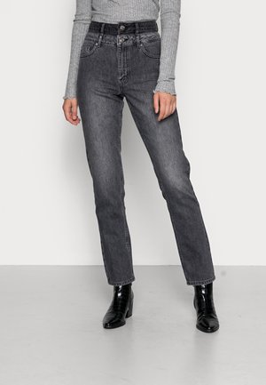 Femme portant un jean noir délavé taille haute avec fermeture à double bouton, un haut gris côtelé à manches longues et des bottines noires à talons blocs.
