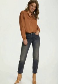 Blusa de cor ferrugem com abertura frontal e mangas franzidas, combinada com jeans azul escuro e botas de salto alto bege. Textura suave.