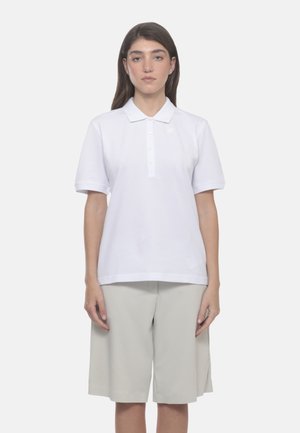 Donna in piedi, rivolta verso l'obiettivo, che indossa una polo bianca a maniche corte e pantaloncini beige chiaro fino al ginocchio su sfondo uniforme.