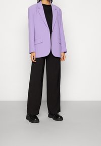 Fioletowa oversize marynarka z wyraźnymi ramionami, zapinana na jeden guzik, z dwiema przednimi kieszeniami, w połączeniu z luźnymi czarnymi spodniami i masywnymi czarnymi botkami.