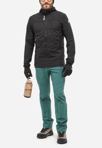 Homme portant une veste noire, un pantalon turquoise, des gants, des chaussures de randonnée, tenant une bouteille d'eau, se tenant devant un fond blanc uni.