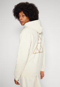Creme hoodie lavet af blødt stof, med et stort beige trekantet logo på ryggen. Designet med en hætte med snoretræk og ribbede manchetter.