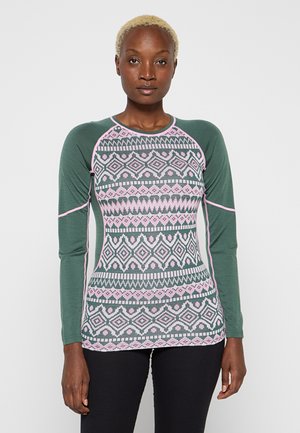 Donna con capelli biondi corti che indossa un maglione a maniche lunghe verde e rosa con motivo e pantaloni neri, in piedi contro uno sfondo semplice.