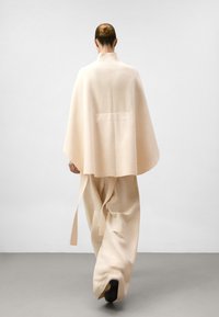 Beige wollen poncho met een hoge kraag, een losse fit en zijbindstrikken, gecombineerd met wijduitlopende bijpassende broeken. Soepele textuur en minimaal ontwerp.