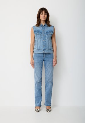 Frau trägt eine ärmellose blaue Jeansweste, passende Jeans mit geraden Beinen und schwarz-weiß gestreifte, spitze Schuhe im Zebra-Design, steht vor einem schlichten Hintergrund.