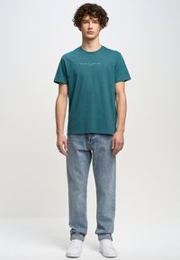 Camiseta de manga corta color aguamarina con un detalle de texto impreso en la parte delantera, combinada con jeans de mezclilla azul claro y zapatillas blancas.