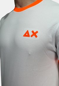 Camicetta bianca con colletto arancione e logo che presenta un triangolo e una X. Realizzata in tessuto testurizzato, aderente al corpo.
