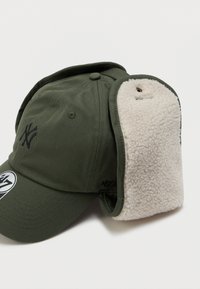 Zelena kapa s mekim, belim umetnim krznom na ušima. Ima zakrivljeni vizir, izšito logotip in nastavljiv trak na hrbtu.