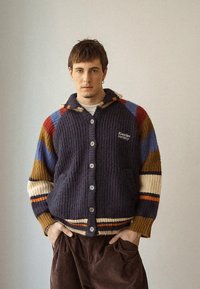 Young man wearing a multicolored knit cardigan with button front and hands in brown corduroy pants pockets, standing against a plain wall.