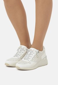 Zapatilla blanca con diseño perforado, acentos plateados y cordones. Presenta un zipper lateral y suela de goma para tracción.