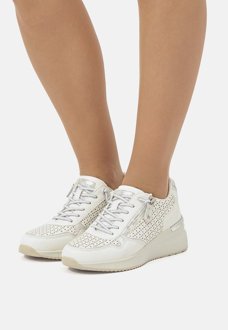 Zapatilla blanca con diseño perforado, acentos plateados y cordones. Presenta un zipper lateral y suela de goma para tracción.