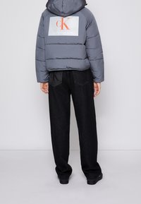 Giacca a vento grigia con cappuccio e grande logo sul retro; abbinata a jeans in denim neri larghi con un logo cucito.