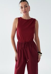Combinaison bordeaux avec un corsage texturé sans manches, taille à cordon de serrage et jambes larges ; dispose d'accents en métal doré et d'un tissu lisse.