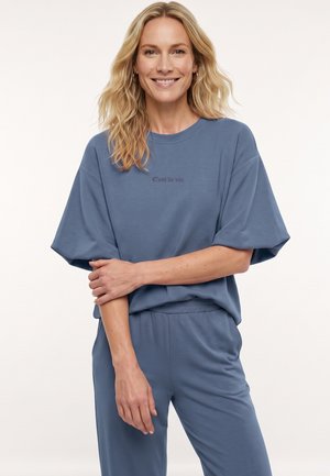 Lachende vrouw met golvend blond haar draagt een losse blauwe bijpassende T-shirt en broek, staand met één arm gekruist over haar middel.