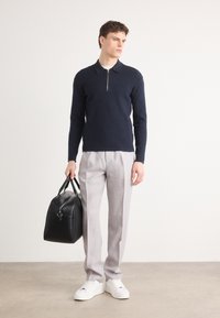 Mornarska polo majica z zadrgo, svetlo sivi krojeni hlače, črna usnjena torba in bele superge; eleganten, modernen dizajn z minimalnimi detajli.
