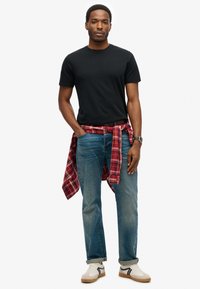 Zwarte t-shirt met ronde hals, blauwe spijkerbroek met een lichte vervaging, rood en zwart geblokt shirt geknoopt om de taille, witte sneakers met bruine accenten.