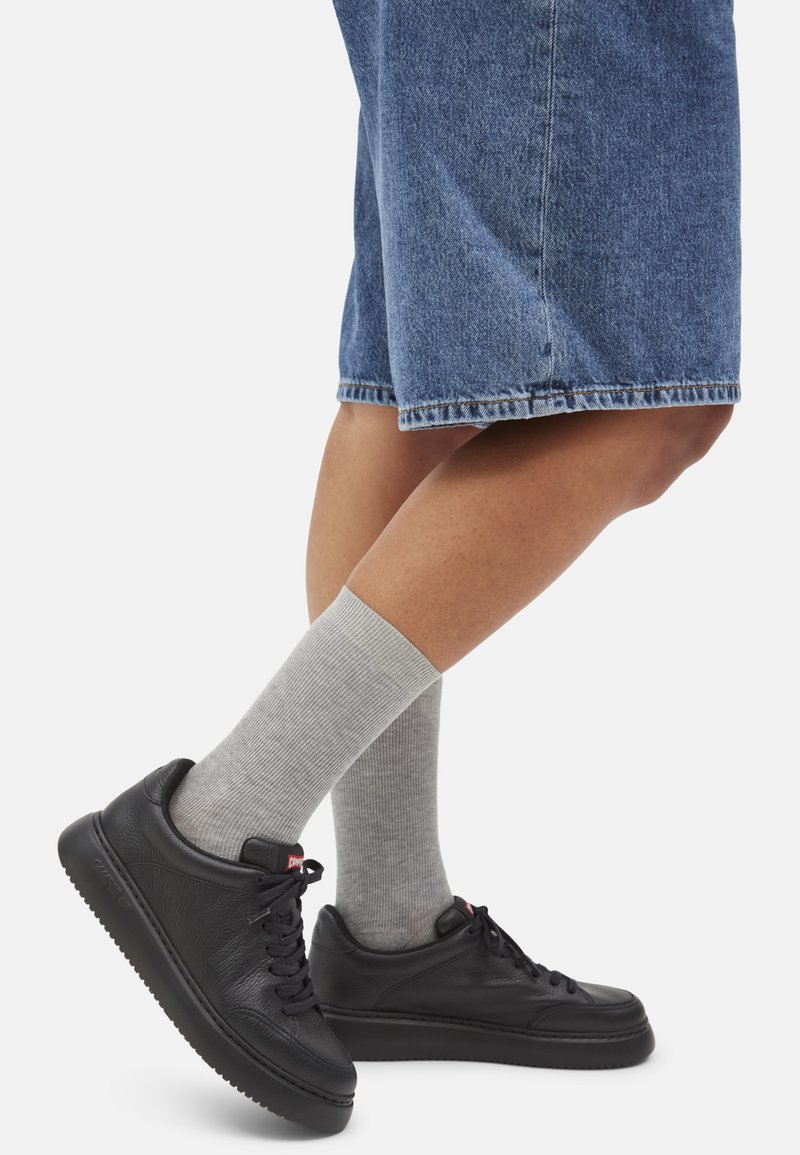 Sneaker nere in pelle con finitura texturizzata, suole in gomma e accenti rossi, abbinate a calze grigie a coste di lunghezza medio e pantaloni short di jeans.
