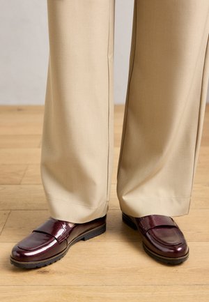 Osoba w beżowych szerokich spodniach i błyszczących, ciemnoczerwonych mokasynach stoi na drewnianej podłodze.