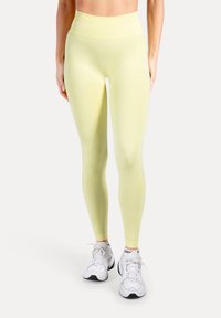 Geel leggings met een hoge taille en een gladde textuur, met een wit merklogo op de tailleband, gecombineerd met witte sportschoenen.