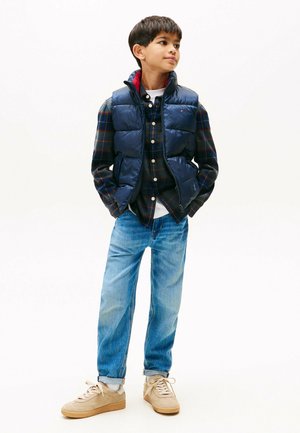 Blaues Steppwesten mit rotem Innenfutter, über einem karierten Hemd getragen. Hellblaue Jeans und beigefarbene Sneaker. Glatte Textur und lässiges Design.