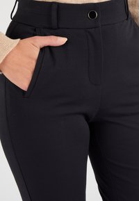 Pantalones negros ajustados con una textura lisa, que presentan bolsillos laterales, un frente plano y un detalle decorativo de botón en tono dorado.