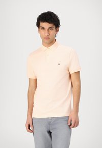 Lys ferskenfarvet bomulds polo-shirt med korte ærmer, en krave og et lille logo på brystet. Båret med lysegrå jeans.
