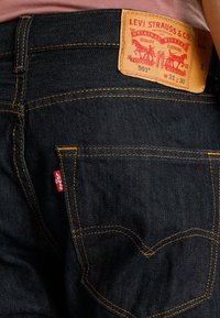 Jeans in denim blu scuro con cuciture arancioni, dotati di un'etichetta in pelle marrone e una linguetta rossa nella tasca posteriore. Dettagli di design rivettati.
