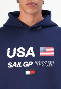 Felpa blu navy con testo "USA" e "SAIL GP TEAM", grafica della bandiera americana e logo di Tommy Hilfiger. Materiale in cotone.
