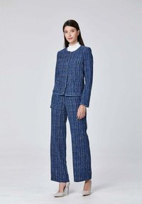 Costume en tweed bleu et blanc composé d'une veste ajustée avec des boutons dorés et d'un pantalon à jambe large, associé à un col roulé blanc et des talons de couleur claire.
