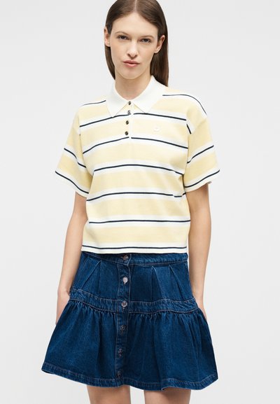 Γυναίκα που φοράει κίτρινη και λευκή ριγέ πόλο μπλούζα με κουμπιά και σκούρα μπλε τζιν φούστα με κουμπιά, στέκεται μπροστά σε απλό φόντο.
