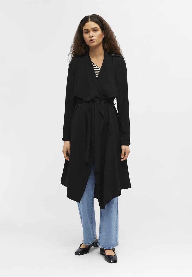 Black long coat with an open front, tied at the waist, featuring long sleeves and a flowing hem, paired with light blue wide-leg jeans and black flats.