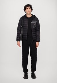 Sort pufferjakke med hætte, prydet med et diagonalt hvidt linjemønster. Kombineret med sorte sweatpants med sideskrevne detaljer og sorte sko.