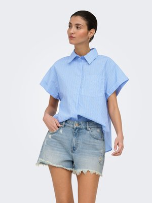 Femme portant une chemise bleue à rayures, à manches courtes et ample, rentrée dans un short en jean clair déchiré avec des ourlets effilochés, debout devant un fond uni.