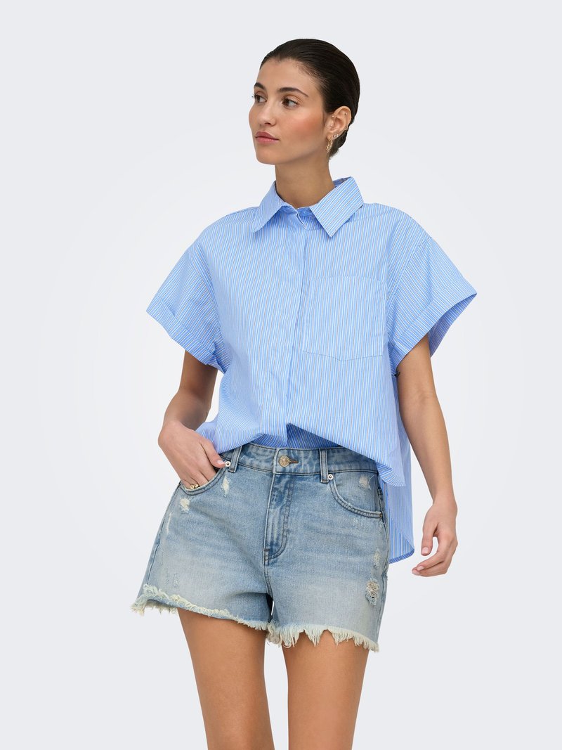 Femme portant une chemise bleue à rayures, à manches courtes et ample, rentrée dans un short en jean clair déchiré avec des ourlets effilochés, debout devant un fond uni.