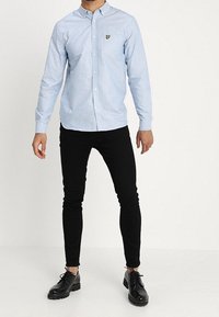 Camisa de manga larga azul claro con botones, cuello y pequeño emblema amarillo, combinada con jeans ajustados negros y zapatos negros con cordones.